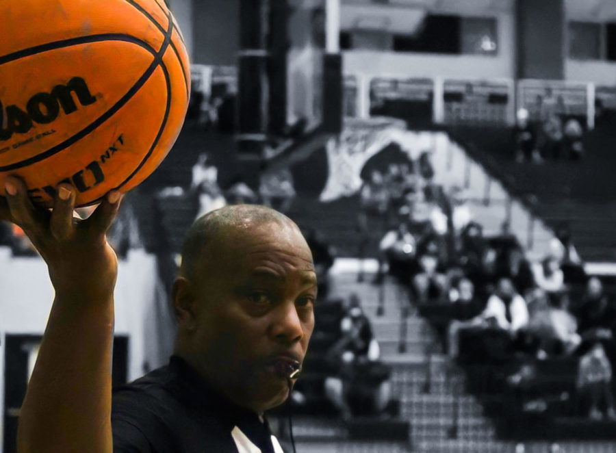 A referee blows his whistle while holding up a basketball during a varsity basketball game.